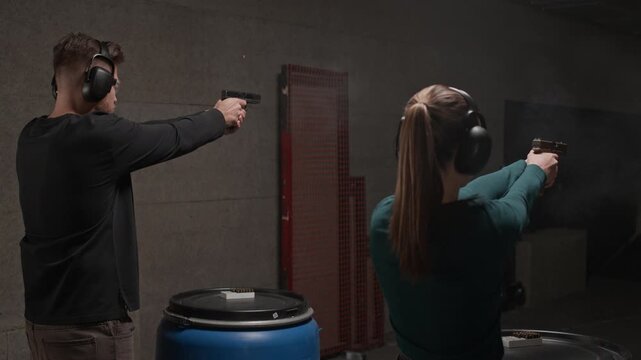 Back view of two people wearing earmuffs and goggles practicing shooting from guns at indoor range