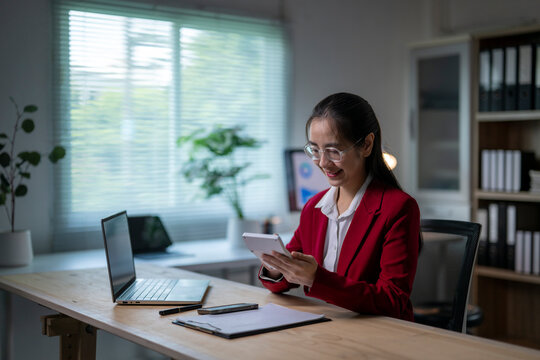 woman in a red suit is sitting at a desk with a laptop and a tablet