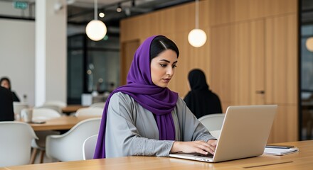 A young woman in a purple hijab works diligently on her laptop in a modern, bright space.