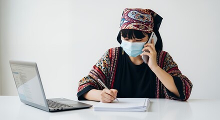 A woman in traditional headwear works from home, using a laptop and phone while wearing a face mask.