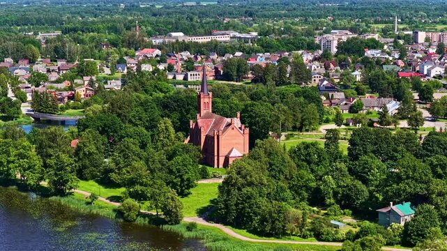 Slow aerial parallax movement reveals Biržai Evangelical Reformed Church nestled in lush greenery and parkland in Lithuania.
.