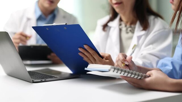 Professor is teaching medical intern students. Doctors and nurses in white coats gather around a table, reviewing patient information on laptops, taking notes during a meeting.