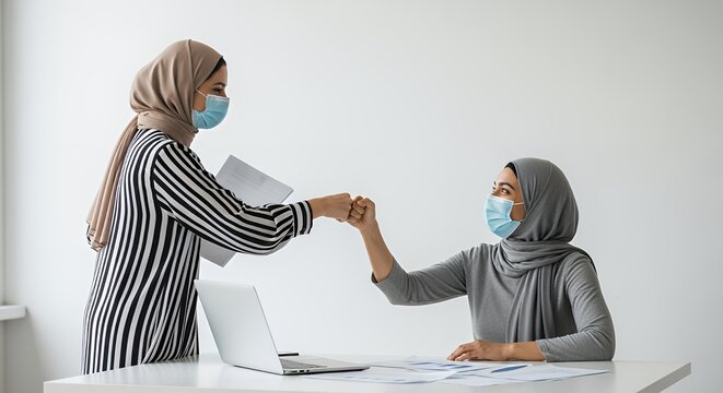 Two Muslim women wearing face masks bump fists in a modern setting, showcasing professional collaboration and safety precautions.