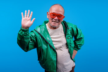 Older Asian Male Wear Green Jacket Waving Hello To Camera Isolated On Blue Background