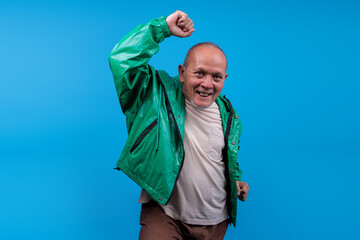 Older Asian Male In Green Jacket Expressing Excitement And Victory Isolated On Blue Background