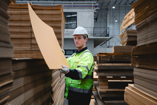 Engineer wearing safety helmet and reflective jacket inspects large sheet of corrugated cardboard inside paper manufacturing plant.
