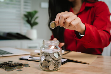 A smiling young woman sits at a desk indoors, holding a piggybank and coins, planning her finances with a calculator, symbolizing saving, security, and financial growth.