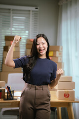Cheerful young woman celebrates success in her office surrounded by packaging boxes representing success in online business, entrepreneurship and e-commerce fulfillment.