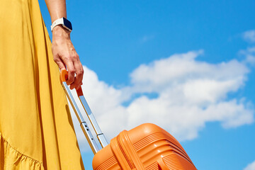 Woman in yellow dress holding an orange suitcase against blue sky with clouds. Close up of female...