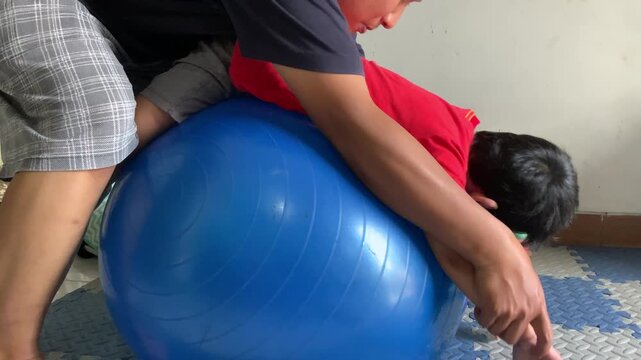 A father assists his son during a home physiotherapy session using a blue therapy ball.