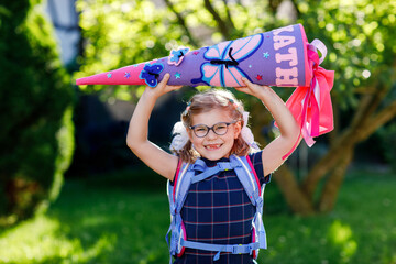 little girl with glasses and a blue backpack, dressed in a navy uniform, holds a colorful school cone called Schultuete and backpack. Happy child smiling, ready for her first day of school