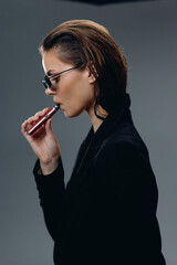 Obraz premium Stylish woman vaping with sunglasses wearing black jacket in studio against grey background