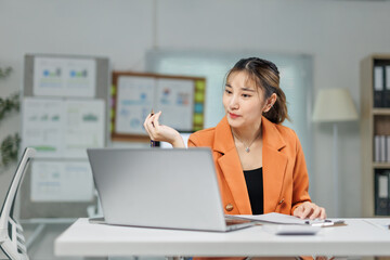 Young Asian businesswoman wearing an orange jacket sitting at her desk in a modern office, gesturing with a pen while engaged in a video call on her laptop