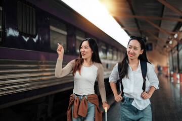 Two young female tourists are walking in a train station, one of them is pointing her finger while pulling a trolley suitcase and the other is carrying a backpack
