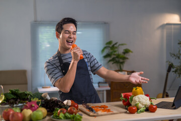 Young man wearing apron singing with carrot like microphone in kitchen while preparing healthy meal with fresh organic vegetables and tablet on table, concept of happy lifestyle and healthy food