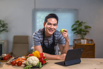 Young man wearing apron holding a green apple and using a tablet in a kitchen with fresh vegetables and fruits on table, promoting healthy eating and technology in cooking
