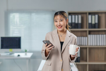 Obraz premium Young asian businesswoman is holding a coffee mug and using a smartphone, standing in a modern office with a desk, computer and bookcase in the background