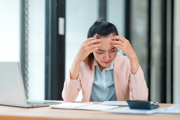 Young Asian businesswoman holding a pen and touching her head with hands, having a headache and feeling tired from working on a laptop computer in the office