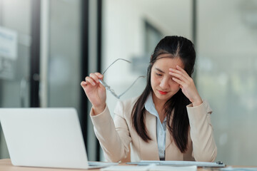 Young Asian businesswoman holding glasses and touching forehead while working on laptop computer at office desk, feeling tired, exhausted, having headache or migraine from overworked or burnout