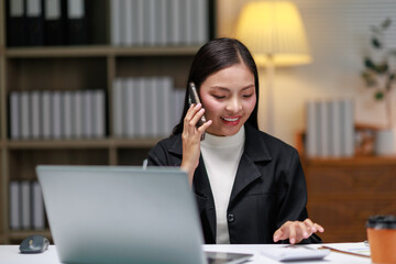 Young professional woman engaged in a phone call and using a calculator while working on a laptop...