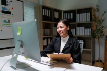 Young happy Asian businesswoman working with desktop computer and holding clipboard, smiling and looking at computer screen in modern office in the evening