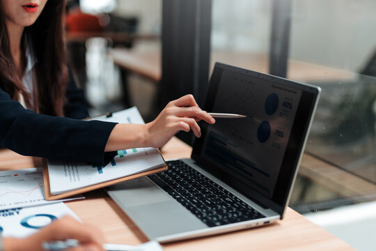 Businesswoman using a pen to point at laptop screen showing financial charts during a meeting with colleagues, analyzing data and discussing strategy