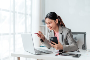 Young Asian businesswoman holding a smartphone while pointing at a laptop screen, collaborating on a project in a bright, modern office filled with natural light