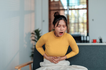 Asian woman experiencing severe abdominal pain while sitting on sofa in living room, showing...