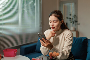 Young Asian woman sitting at a table in her living room, wearing a facial mask and applying makeup...