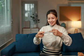 Happy young Asian woman sitting on a blue sofa in her living room at night, holding a moisturizing...