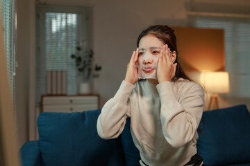 Young woman sitting comfortably on a sofa in her living room, applying a cosmetic face mask and...