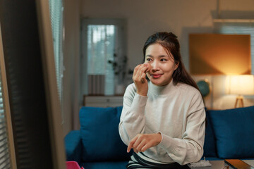 Sitting comfortably on her sofa in the living room, a young Asian woman is meticulously applying...