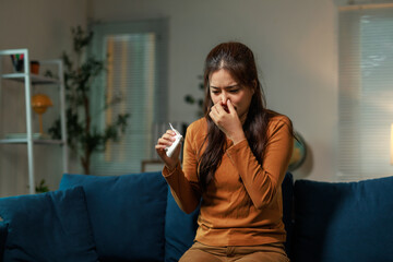 Sick young woman sitting on a sofa in a cozy living room at night, holding a nasal spray and...
