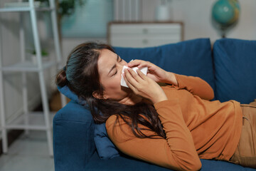 Sick young woman lying on the sofa, blowing her nose while battling cold or allergy symptoms and...