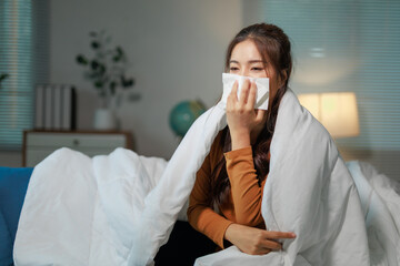 Sick young asian woman wrapped in blanket is blowing her nose while sitting on sofa in living room...