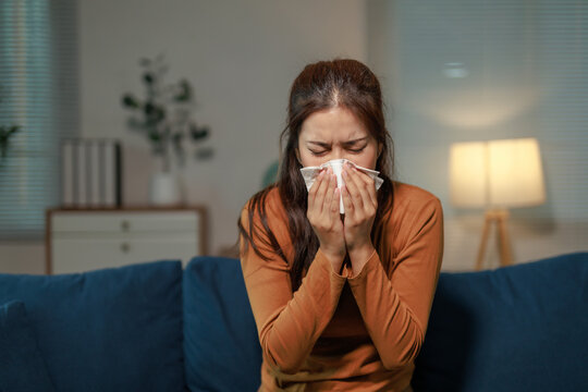Young asian woman is blowing her nose with a tissue while sitting on a sofa in the living room at night, suffering from a cold or allergies