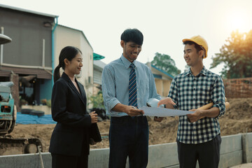 Asian architect holding blueprint and explaining building plan to businesswoman and construction...