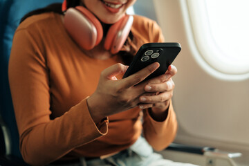Happy tourist enjoying a flight while using a mobile phone, wearing headphones and smiling, engaged in browsing the internet or connecting on social media