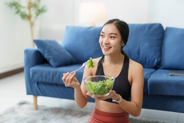 Happy asian sportswoman smiling while eating a healthy salad in her living room after a workout session, promoting a healthy lifestyle and post exercise nutrition