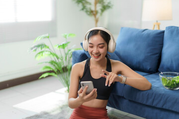 Asian woman wearing headphones and sportswear, sitting on the floor with a smartphone, enjoying...