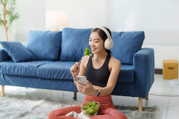 Happy Asian sportswoman enjoying leisure time in a modern living room, wearing headphones, eating a...