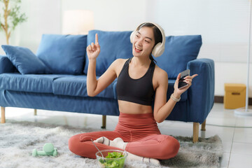 Happy asian girl in sportswear sitting on carpet in living room listening to music with headphones...