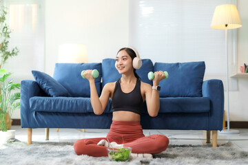 Happy athletic woman wearing wireless headphones lifts light dumbbells in the lotus position on a...