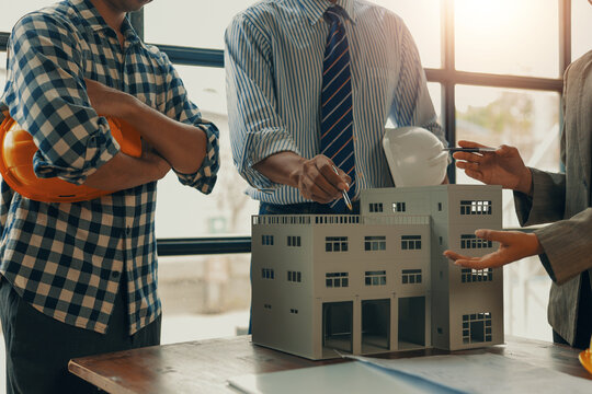 Architects and engineers engaging in a discussion over a building model during a meeting in the office, pointing at the details of the model while holding safety helmets