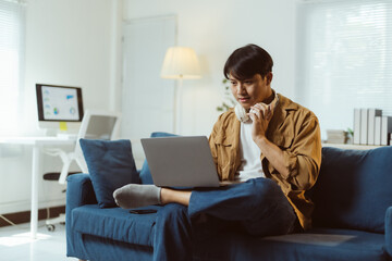 Young Asian man sitting comfortably on a sofa in his living room, working from home while using a...