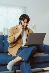 Young Asian man sitting comfortably on the sofa in his living room, working remotely on his laptop while engaged in a phone conversation, exuding focus and productivity