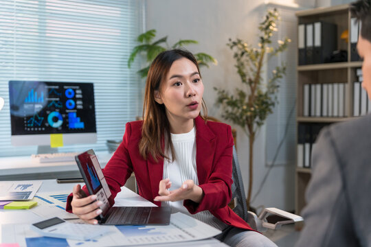 Asian businesswoman showing infographic on tablet and explaining project to colleague during business meeting in office, explaining charts and graphs using laptop at work