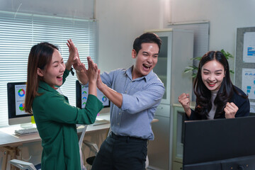 Euphoric Asian businesspeople celebrating a successful project, giving high fives while gathered around a computer in the office, radiating joy and teamwork in their workplace