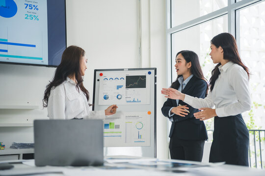 Businesswomen analyzing financial statistics displayed on whiteboard and discussing marketing strategy during corporate meeting in modern office