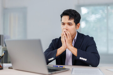 Asian businessman covering his mouth with his hands while looking with concern at his laptop,...